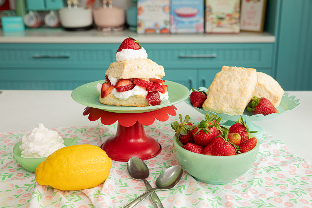 Lemon Strawberry Shortcake on a green plate with a bowl of fresh strawberries beside it and a lemon on the table.