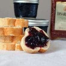 A stack of toast bread slices, one with boozy sweet cherry preserves spread over it. Small jar containers with cherry preserved on the background.