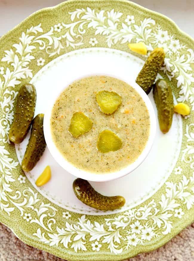 top down shot of Dill Pickle Soup numa pequena assadeira redonda branca com alguns pickles de endro à volta de um prato branco pronto a ser apreciado!