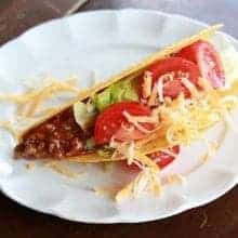 Close up of Classic Weeknight Tacos with shredded lettuce and beef mixture, garnish with grated cheddar, sour cream, tomatoes and green onions