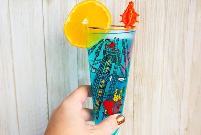 woman's hand holding a blue AMF cocktail drink garnished with an orange slice that's in a vintage pirates motif glass