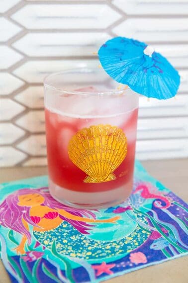 Bay Breeze Cocktail in a glass emblazoned with a gold shell and garnished with a small umbrella on a mermaid napkin