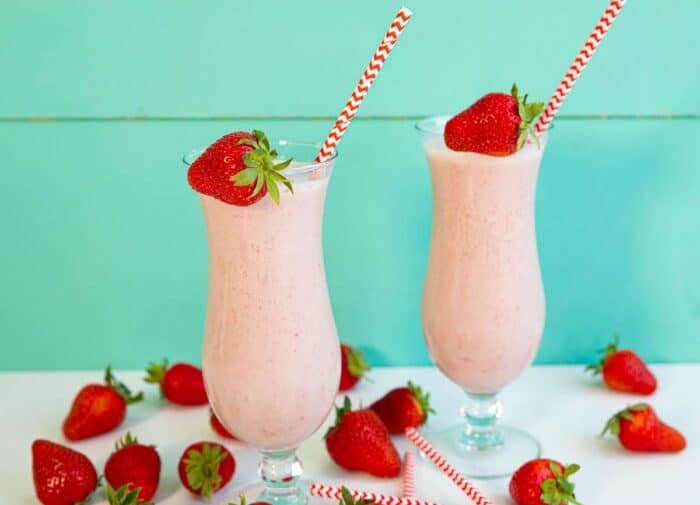 Wider shot of two strawberry milks with loose and strawberries and white and red straws surrounding them.