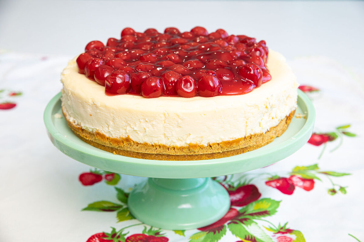 a whole Philadelphia cheesecake topped with cherry pie filling on a Jadeite cake stand