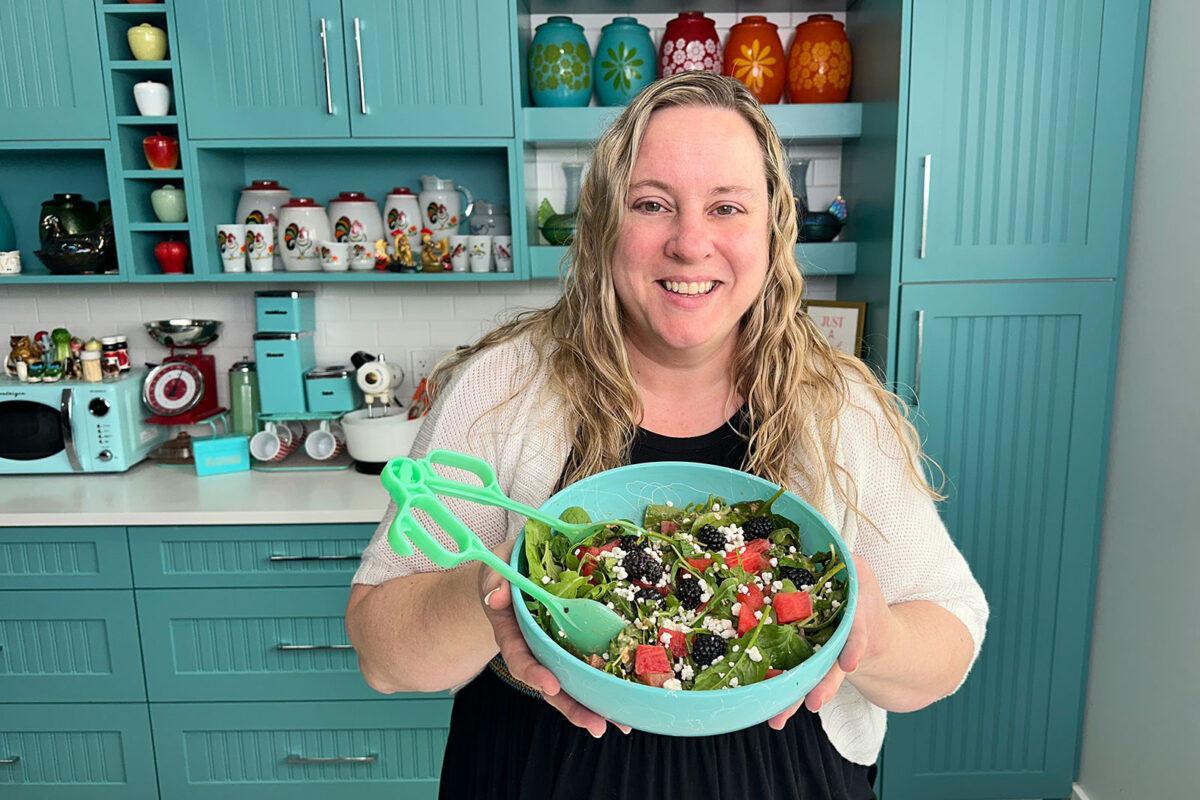 Karlynn holing a bowl of watermelon salad with feta and mint in a green bowl with green tongs. 