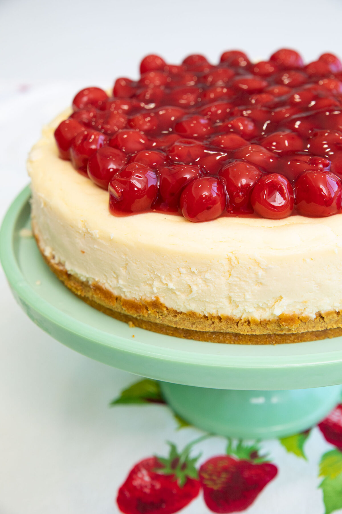 close up photo of a whole Philadelphia cheesecake topped with cherry pie filling on a Jadeite cake stand