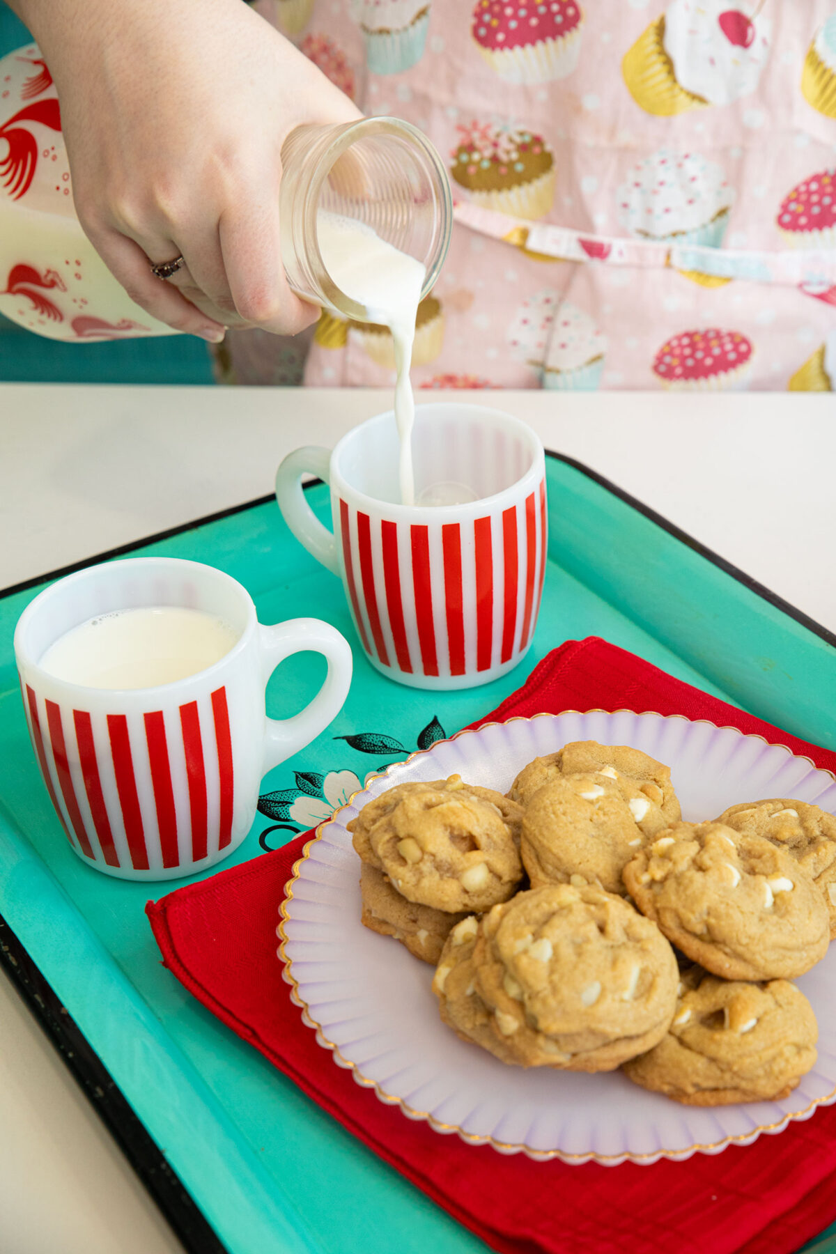 white chocolate chip macadamia nut cookies on a white plate with two cups of milk on a teal tray.