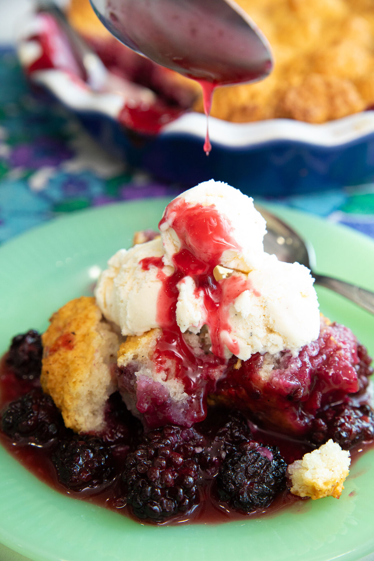 blackberry cobbler on a green plate topped with ice cream and a spoon dripping sauce onto the top.